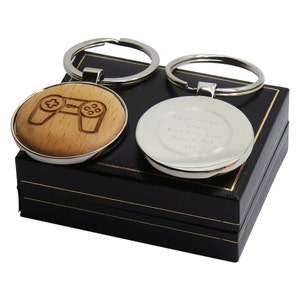 以下が含まれることがあります： 木製の円形にレーザー彫刻されたビデオゲームコントローラーが付いたシルバーのキーチェーン。キーチェーンの反対側は、シルバーの円形に「Happy Birthday Josh Love from Mum & Dad」というメッセージが刻まれています。キーチェーンは黒いギフトボックスに入っています。