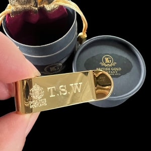 May include: Gold-coloured bottle opener with the inscription "T.S.W." and the Police Scotland logo. The bottle opener is in a black velvet pouch with a gold-coloured drawstring.