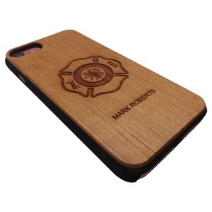Pode incluir: Uma capa de iPhone de madeira com uma borda preta. A capa apresenta um distintivo de bombeiro gravado a laser com a palavra "Fire" e um machado e uma escada cruzados. O nome "MARK ROBERTS" está gravado abaixo do distintivo.