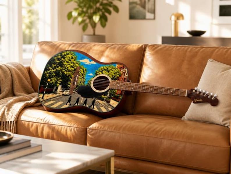 Puede incluir: Guitarra ac&uacute;stica con una imagen colorida de Abbey Road en el cuerpo, apoyada en un sof&aacute; de cuero marr&oacute;n. La guitarra tiene un m&aacute;stil y clavijero marrones, y un logotipo de Fender. Una almohada y una manta beiges tambi&eacute;n est&aacute;n en el sof&aacute;.