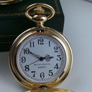 May include: A gold pocket watch with a white face and black numbers. The watch has a chain attached to it and says "BRITISH GOLD Co QUARTZ" on the face.