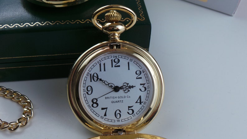 May include: A gold pocket watch with a white face and black numbers. The watch has a chain attached to it and says "BRITISH GOLD Co QUARTZ" on the face.
