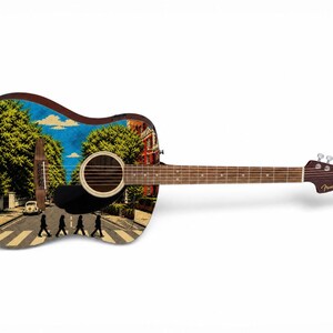 Puede incluir: Guitarra ac&uacute;stica con un dise&ntilde;o personalizado que presenta la ic&oacute;nica escena de Abbey Road. La guitarra tiene un cuerpo marr&oacute;n, una boca negra y un clavijero Fender. La imagen muestra un d&iacute;a soleado con &aacute;rboles y edificios.