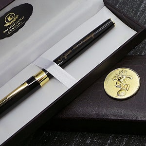 Peut inclure: Un stylo noir avec des accents dorés et une inscription personnalisée dans une boîte brune. Le stylo est dans une boîte blanche avec un logo doré qui indique "British Gold Company Exclusive Gift Gifts".