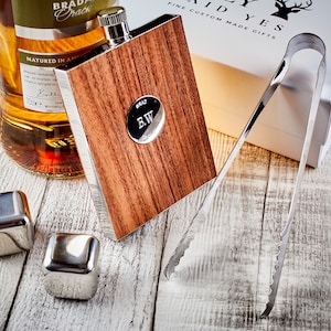 May include: A wooden and stainless steel hip flask with a personalised engraved plate that says "B.W.". The flask is next to a bottle of whisky, a set of stainless steel ice cubes, and a pair of tongs.