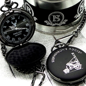 May include: Black pocket watch with a silver chain and a black and silver pocket watch case. The watch face has Roman numerals and a stag design. The case is engraved with "Stephenson's Rocket".