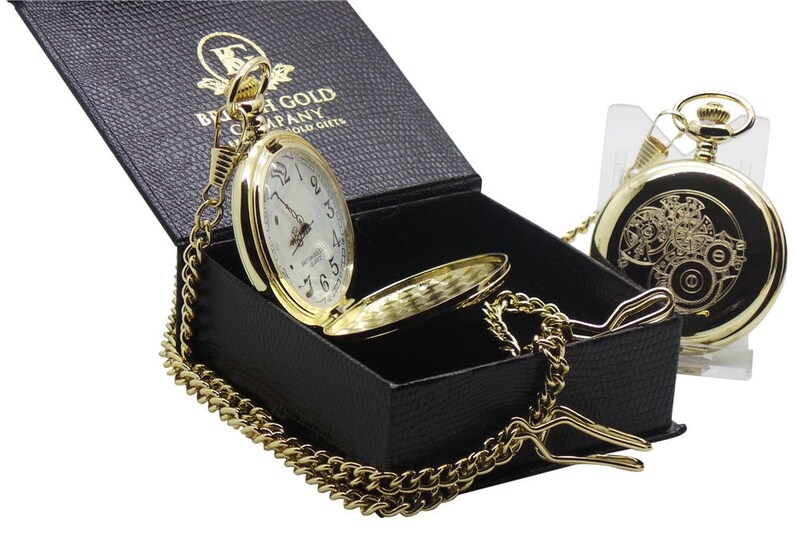 May include: A gold pocket watch with a white face and black Roman numeral hour markers. The watch is in a black box with a gold chain. The box has the text "British Gold Company" on it.
