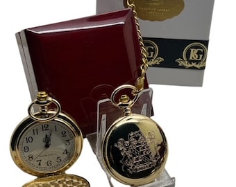 Puede incluir: Dos relojes de bolsillo dorados, uno abierto para revelar la esfera del reloj, el otro cerrado con un diseño ornamentado. También se ven una cadena dorada y una caja blanca con detalles dorados. Los relojes se exhiben en un soporte transparente.