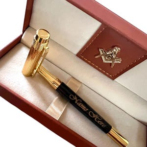 Pode incluir: Uma caneta dourada com um corpo preto, apresentada numa caixa vermelha com interior castanho. A caneta tem o texto "Name Here" gravado no corpo. A caixa também apresenta um emblema maçónico.