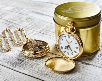 Puede incluir: Un reloj de bolsillo dorado con una esfera blanca y números romanos. El reloj está abierto y la parte posterior está grabada con un escudo de armas. El reloj está en una lata dorada con el texto "ANY NAME" grabado en la tapa.
