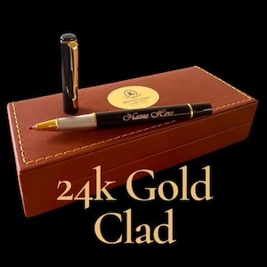 Può includere: Una penna nera e oro con la scritta "Name Here" sul fusto, appoggiata su una scatola di pelle marrone. Il cappuccio della penna è di lato. La scatola ha un sigillo d'oro con la scritta "British Gold Company". Il testo "24k Gold Clad" è in basso.