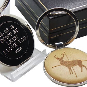 Pode incluir: Um chaveiro de prata com uma etiqueta redonda gravada que diz "12-06-18 Não se atrase! Eu te amo XXX". O chaveiro também tem uma etiqueta redonda de madeira com uma imagem gravada a laser de um veado com uma mira.