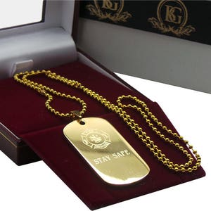May include: A gold-coloured necklace with a dog tag pendant. The pendant features a firefighter emblem and the words "Stay Safe" engraved on it. The necklace is presented in a red velvet box.
