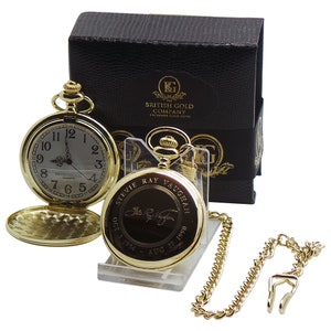 Könnte beinhalten: Zwei goldfarbene Taschenuhren, eine offen und eine geschlossen, mit passender Kette und Verschluss. Die geschlossene Uhr zeigt den Namen "Stevie Ray Vaughan" und Daten. Eine schwarze Schachtel mit goldfarbenen Buchstaben befindet sich im Hintergrund.