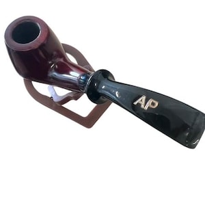 May include: A dark red wooden pipe with a black mouthpiece and a gold "AP" logo. The pipe is resting on a wooden stand.
