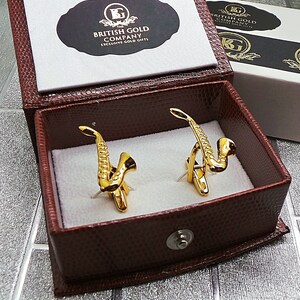 Pode incluir: Um par de botões de punho de saxofone dourados em uma caixa de couro marrom. A caixa tem um rótulo branco com o texto "The British Gold Company Exclusive Gold Gifts".