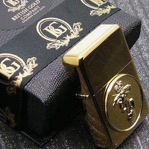 Peut inclure: Un briquet doré avec une boîte noire et dorée. La boîte porte l'inscription "The British Gold Company Exclusive Gold Gifts". Le briquet est orné d'un motif doré avec une couronne et une figure.
