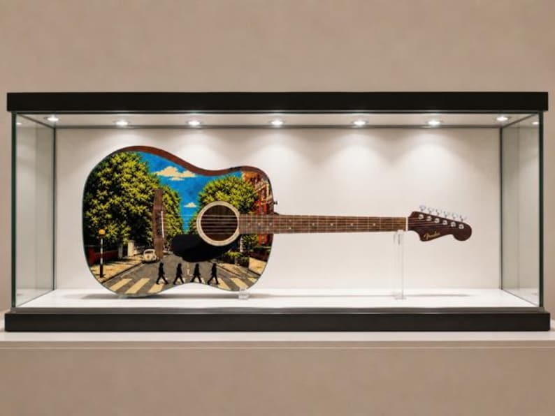 Puede incluir: Guitarra ac&uacute;stica con un dise&ntilde;o personalizado que presenta la ic&oacute;nica escena del cruce de Abbey Road. La guitarra se exhibe en una vitrina de cristal, mostrando la colorida obra de arte en el cuerpo. La guitarra tiene un m&aacute;stil y clavijero marrones.