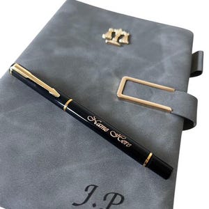 Puede incluir: Un diario de cuero gris con un bolígrafo dorado y un emblema de balanza dorado. El bolígrafo negro tiene detalles dorados y las palabras "Name Here" están escritas en él. Las iniciales "J.P" están grabadas en la portada del diario.