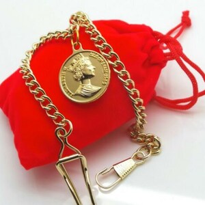 May include: A gold pocket watch chain with a round gold pendant featuring a profile of a woman with a crown. The chain is on a red velvet pouch.