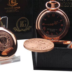 May include: A rose gold pocket watch with a black face and Roman numeral hour markers. The watch is in a black box with a rose gold coin and a rose gold pocket watch with a Bitcoin logo on the face.