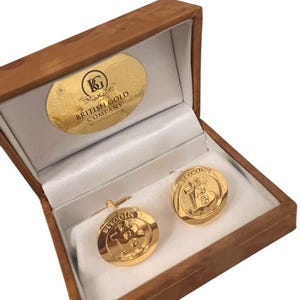 May include: Gold cufflinks in a wooden box with a white interior. The cufflinks feature a gold Bitcoin logo with the text "Bitcoin" above it. The box has a label that reads "The British Gold Company".