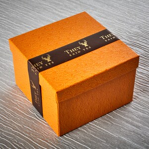 Op de afbeelding: Een oranje geschenkdoos met een bruin lint waarop "They Said Yes" staat met een gouden hertenlogo.