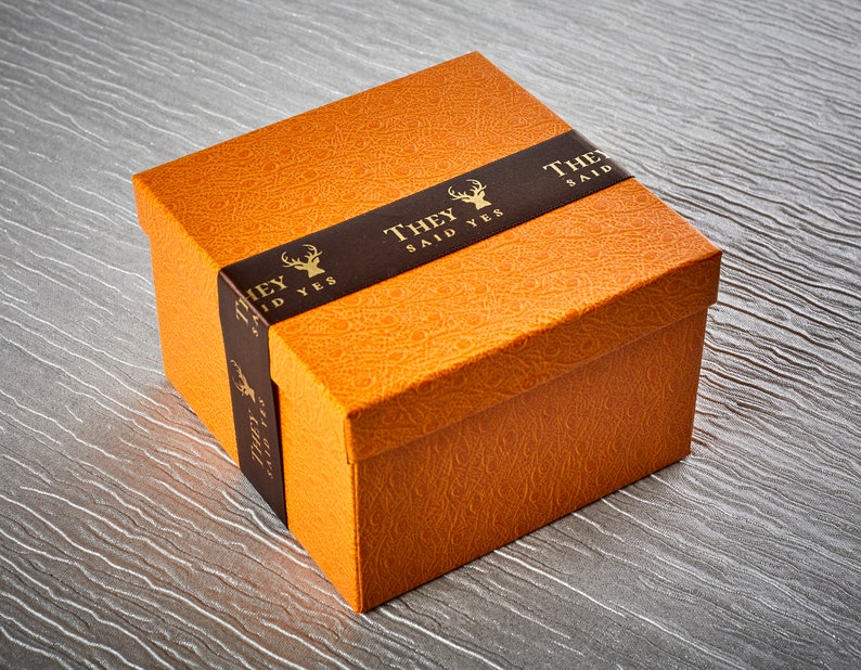 Op de afbeelding: Een oranje geschenkdoos met een bruin lint waarop "They Said Yes" staat met een gouden hertenlogo.