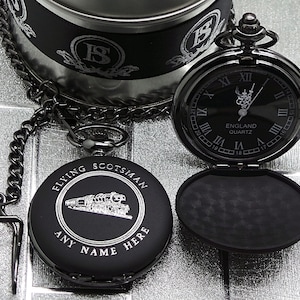 Puede incluir: Reloj de bolsillo negro con una cadena plateada y una inscripción plateada que dice "FLYING SCOTSMAN" y "ANY NAME HERE". La esfera del reloj es negra con marcadores de hora en números romanos plateados y una inscripción plateada que dice "ENGLAND QUARTZ".