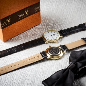 Op de afbeelding: Twee gouden polshorloges met zwarte leren banden, een met een witte wijzerplaat en de andere met een gouden wijzerplaat. De horloges liggen op een wit oppervlak met een zwarte vlinderdas.