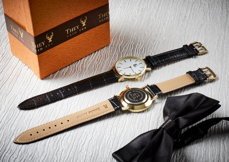 Op de afbeelding: Twee gouden polshorloges met zwarte leren banden, een met een witte wijzerplaat en de andere met een gouden wijzerplaat. De horloges liggen op een wit oppervlak met een zwarte vlinderdas.