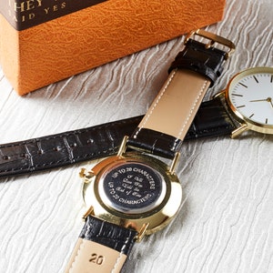 Op de afbeelding: Een gouden polshorloge met een zwarte leren band. De wijzerplaat van het horloge is wit met zwarte wijzers en een zwart logo. De achterkant van het horloge heeft een zwarte gegraveerde plaat met de tekst "Tot 20 tekens Ik zal je tot het einde der tijden liefhebben Tot 20 tekens".
