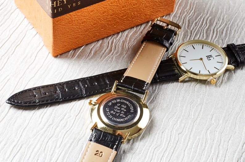 Op de afbeelding: Een gouden polshorloge met een zwarte leren band. De wijzerplaat van het horloge is wit met zwarte wijzers en een zwart logo. De achterkant van het horloge heeft een zwarte gegraveerde plaat met de tekst "Tot 20 tekens Ik zal je tot het einde der tijden liefhebben Tot 20 tekens".