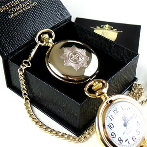 May include: Gold pocket watch with a chain and a black box. The watch face has Roman numerals and a small emblem. The back of the watch has a gold emblem.