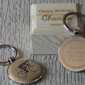 May include: A silver keyring with a round wooden charm featuring a silhouette of a person riding a bicycle. The back of the charm is silver and engraved with the text "Any Name Any Message Here up to 50 Characters". The keyring is packaged in a small wooden box with the text "Happy Birthday Charlie 05/02/1" engraved on the top.
