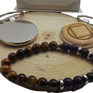 Può includere: Due portachiavi d'argento con ciondoli rotondi in legno, uno con una superficie liscia e l'altro con un design quadrato intagliato. Un braccialetto di perline marroni e viola con perline di occhio di tigre e ametista è esposto su una superficie di legno.
