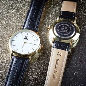 Op de afbeelding: Twee goudkleurige polshorloges met zwarte leren banden. Het horloge aan de linkerkant heeft een witte wijzerplaat met zwarte wijzers en een gouden 'KG'-logo. Het horloge aan de rechterkant heeft een gouden achterkant met een inscriptie die luidt 'Tot 20 tekens Ik zal van je houden tot het einde der tijden Tot 20 tekens'. De band van het horloge aan de rechterkant heeft '20 Echt leer' gegraveerd op de achterkant.