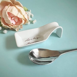 May include: White ceramic spoon rest with a handle, featuring the handwritten text "Love & hugs, Heather".