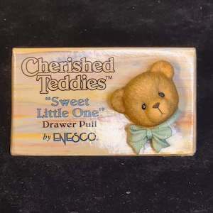 May include: A rectangular drawer pull featuring a brown teddy bear with a light blue bow. The text on the pull reads "Cherished Teddies", "Sweet Little One", and "Drawer Pull by Enesco".