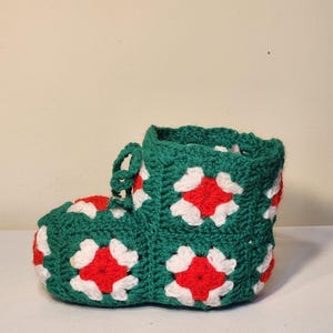 May include: A handmade crochet bootie in green, red, and white. The bootie features a granny square design with red and white flower-like patterns on a green background. The top has a green cuff and a lace closure.