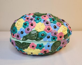 May include: A light green egg-shaped trinket box with a floral design. The box is decorated with painted flowers in shades of pink, yellow, blue, and purple. The flowers have black centres and green leaves.