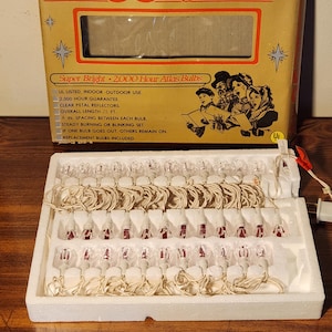 以下が含まれることがあります： 段ボール箱に入ったヴィンテージのミニチュアライトセット。箱はタン色で、赤と金のアクセントがあり、「35 Light Set」と「Golden Sleigh」の文字が特徴です。セットには35個のクリア電球が含まれており、長さは7.62メートルです。