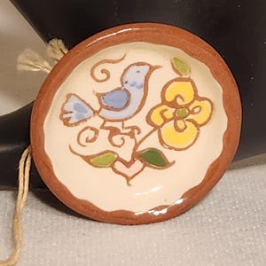 May include: A small, round ceramic dish with a terracotta-colored rim and a cream-colored center. The dish features a hand-painted design of a light blue bird, a yellow flower with green leaves, and a small pink heart, all outlined in brown.