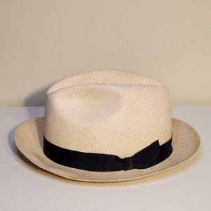 May include: A light beige straw fedora hat featuring a pinched crown and a wide brim. The hat is accented with a solid black grosgrain ribbon band that wraps around the base of the crown, tied in a small bow on the right side of the hat.