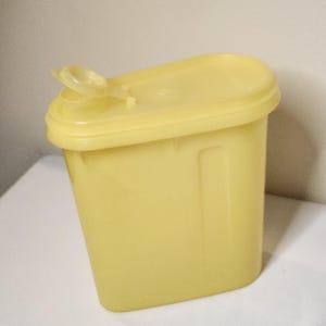May include: A yellow plastic container with a rectangular shape and a lid. The lid has a small, hinged opening. The container is sitting on a white surface.