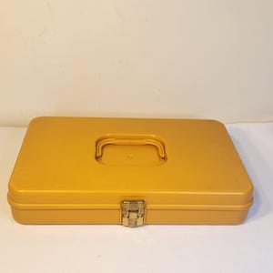 May include: Mustard yellow rectangular case with a handle and a gold-colored clasp. The case is closed and appears to be made of plastic. The handle is centered on the top of the case.