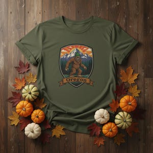 Puede incluir: Camiseta verde oliva con un gráfico de Bigfoot frente a un paisaje montañoso y la palabra "Oregon" en una pancarta. La camiseta está rodeada de hojas de otoño y pequeñas calabazas.