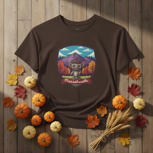 May include: A brown t-shirt featuring a cartoon illustration of a creature with a mountain and forest backdrop. The text "Massachusetts" is displayed on a banner. The shirt is surrounded by pumpkins, leaves, and wheat.