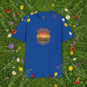 Op de afbeelding: Een koningsblauw T-shirt met een cirkelvormige afbeelding met het woord "NEBRASKA" boven een zonsondergangscène. De woorden "CORN - HUSKER - STATE" staan eronder. Het T-shirt is omgeven door groen gras en kleurrijke bloemen.