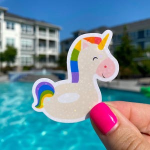 May include: A white unicorn sticker with a rainbow mane, tail, and horn. The sticker has a light beige body with small star-shaped sparkles. The background is a swimming pool and buildings.